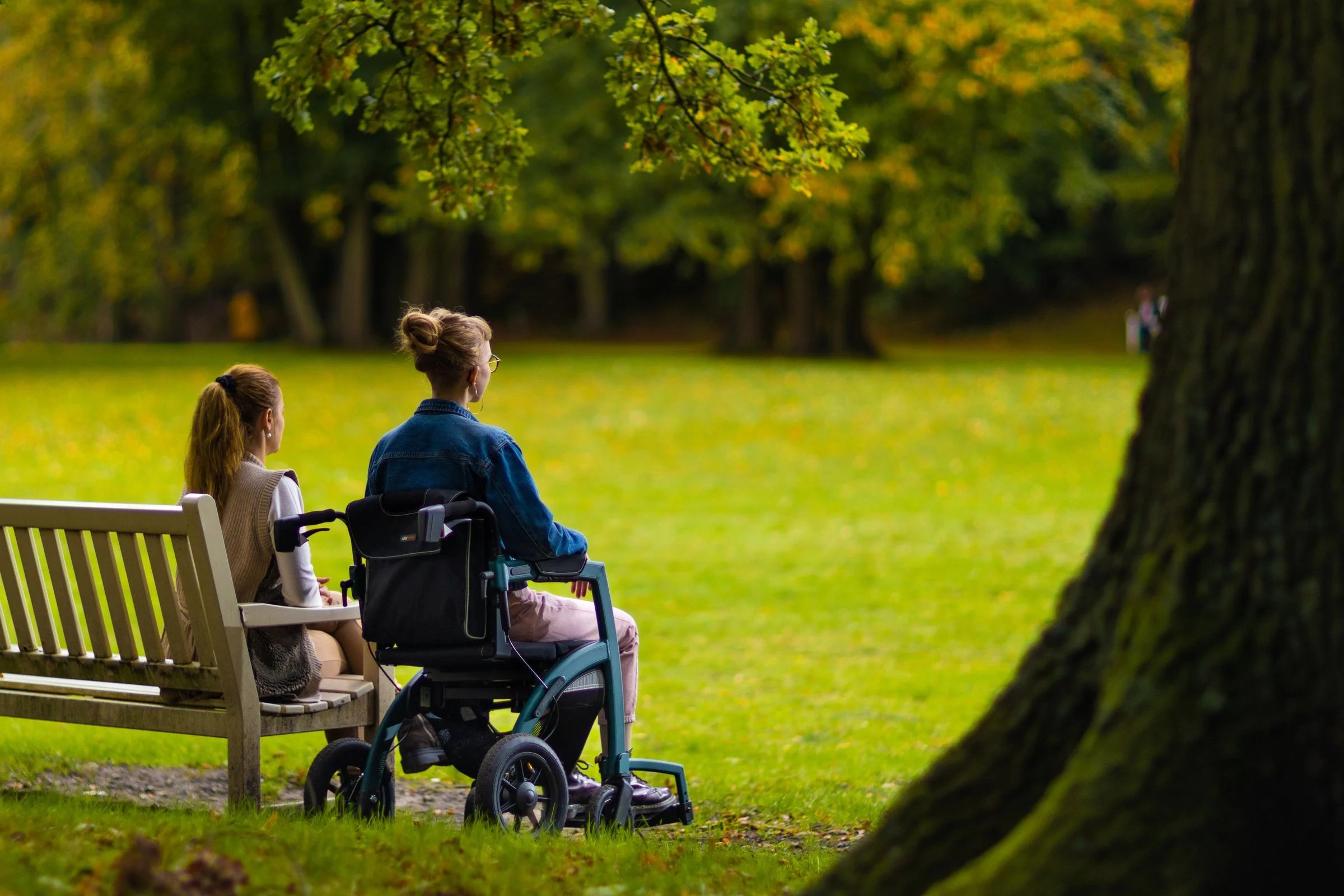 Two women sitting together in a park, one using a mobility chair